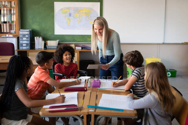 Caucasian female teacher with digital tablet teaching group of multi ethnic kids in the classroom. Primary education social distancing health safety during Covid19 Coronavirus pandemic. — Stock Photo