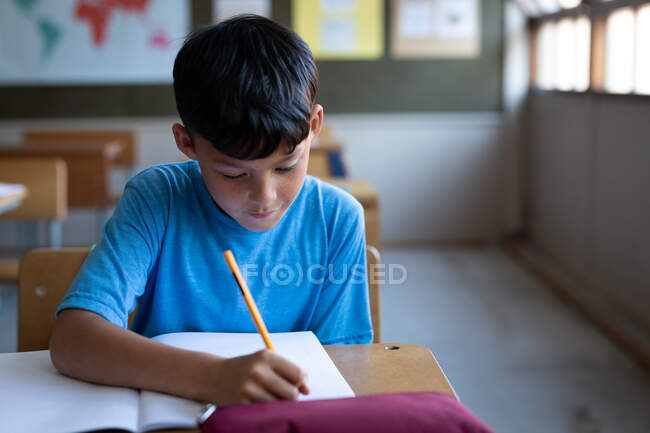 Mixte garçon de race écrit dans un livre tout en étant assis sur son bureau à l'école. Enseignement primaire distanciation sociale sécurité sanitaire pendant la pandémie de coronavirus Covid19. — Photo de stock