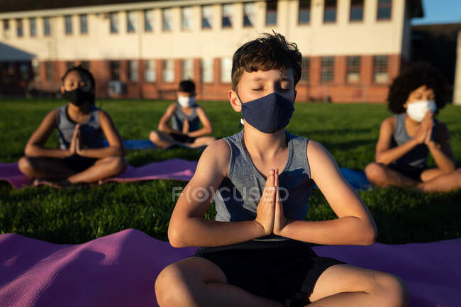 Gruppo di bambini multietnici che indossano maschere facciali che praticano yoga nel giardino della scuola. Istruzione primaria distanza sociale sicurezza sanitaria durante la pandemia di Covid19 Coronavirus. — Foto stock