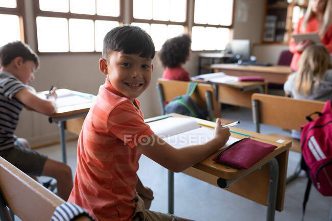 Ritratto di un ragazzo di razza mista sorridente seduto sulla scrivania a scuola. Istruzione primaria distanza sociale sicurezza sanitaria durante la pandemia di Covid19 Coronavirus. — Foto stock