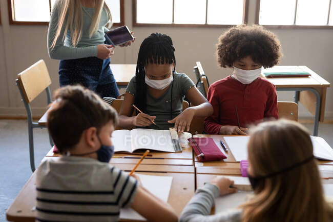 Kaukasische Lehrerin mit Gesichtsmaske, die eine Gruppe multiethnischer Kinder unterrichtet. Grundschulbildung soziale Distanzierung der Gesundheitssicherheit während der Covid19 Coronavirus-Pandemie. — Stockfoto