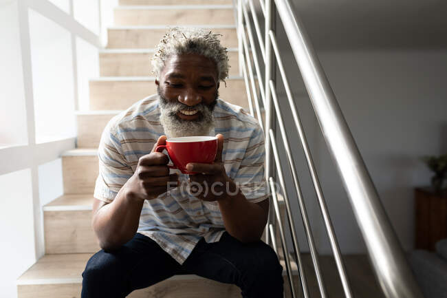 Uomo anziano afroamericano seduto sulle scale, con in mano una tazza di caffè e sorridente, distanza sociale e isolamento in quarantena — Foto stock