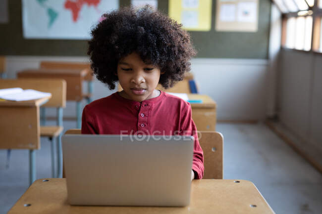 Menino de raça mista usando laptop enquanto sentado em sua mesa na escola. Educação primária distanciamento social segurança sanitária durante Covid19 pandemia de coronavírus. — Fotografia de Stock