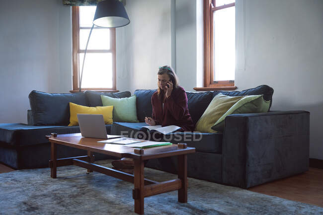 Кавказька жінка, яка проводить час удома, спілкується на відстані і самотності в карантині, сидить на дивані в вітальні, користується ноутбуком, розмовляє по телефону. — стокове фото
