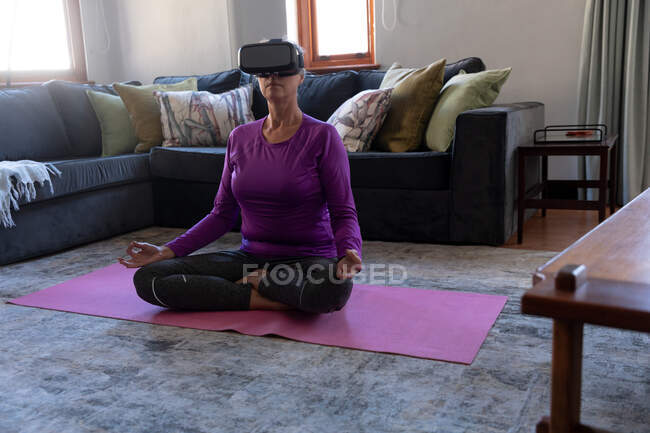 Mujer caucásica disfrutando del tiempo en casa, distanciamiento social y autoaislamiento en cuarentena, ejercicio en la sala de estar, con auriculares VR, sentado en el suelo con las piernas cruzadas, meditando. - foto de stock