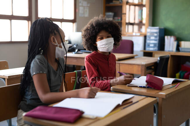 Multi ethnic boy and girl sitting at desks wearing face masks in classroom. Primary education social distancing health safety during Covid19 Coronavirus pandemic. — Stock Photo