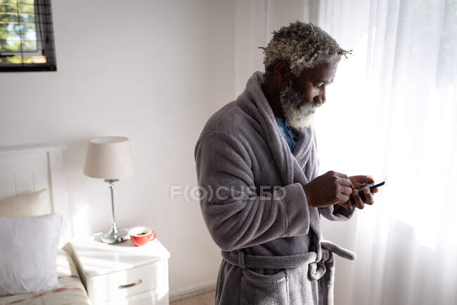 Homme âgé afro-américain debout dans une chambre à coucher, utilisant un smartphone, la distance sociale et l'isolement personnel en quarantaine verrouillage — Photo de stock