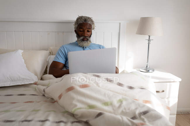Hombre mayor afroamericano acostado en una cama en un dormitorio, usando una computadora portátil y sonriente, distanciamiento social y aislamiento en cuarentena - foto de stock