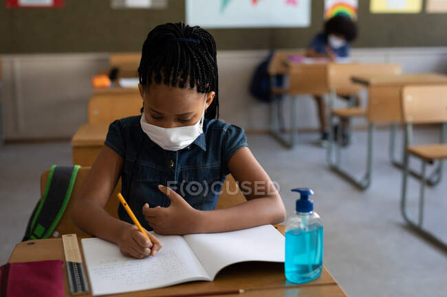 Gemischte Rassenmädchen tragen Gesichtsmaske, während sie am Schreibtisch im Klassenzimmer sitzen. Grundschulbildung soziale Distanzierung der Gesundheitssicherheit während der Covid19 Coronavirus-Pandemie. — Stockfoto