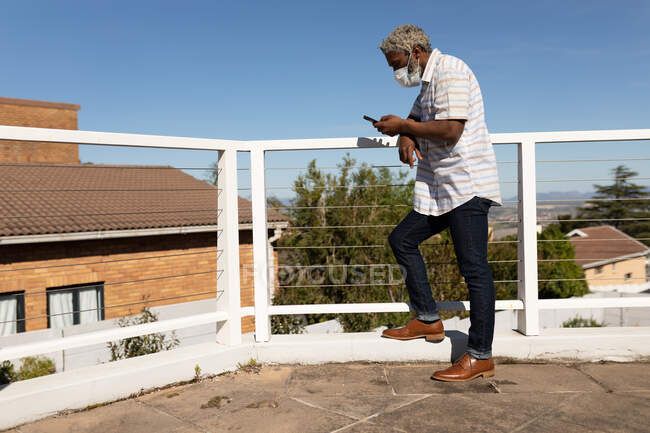 Homme âgé afro-américain debout sur un balcon, en utilisant un smartphone, la distance sociale et l'isolement personnel en quarantaine verrouillage — Photo de stock