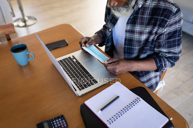 Afro-americano idoso sentado ao lado de uma mesa, usando laptop, segurando uma máscara facial, distanciamento social e auto-isolamento em bloqueio de quarentena — Fotografia de Stock