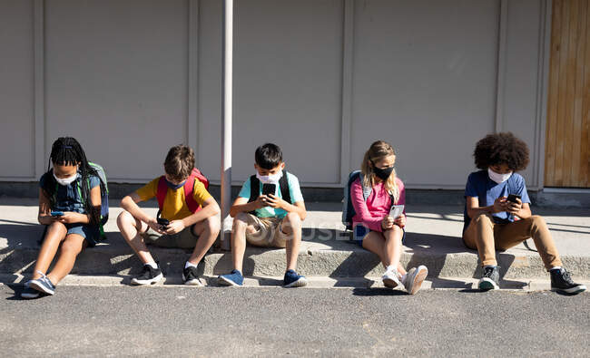 Grupo multiétnico de niños de la escuela primaria que usan máscaras faciales usando teléfonos inteligentes mientras están sentados juntos. Educación primaria distanciamiento social seguridad sanitaria durante la pandemia del Coronavirus Covid19 - foto de stock