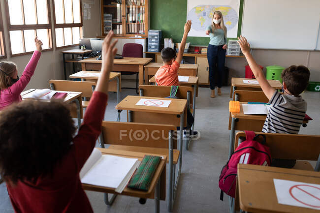 Group of multi ethnic kids sitting on their desk during the lesson with a female teacher wearing a face mask. Primary education social distancing health safety during Covid19 Coronavirus pandemic. — Stock Photo