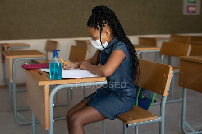 Chica de raza mixta que usa máscara facial mientras está sentada en su escritorio en el aula con un desinfectante. Educación primaria distanciamiento social seguridad sanitaria durante la pandemia del Coronavirus Covid19. - foto de stock