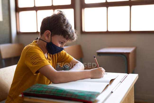 Ragazzo caucasico seduto alla scrivania con la maschera in classe. Istruzione primaria distanza sociale sicurezza sanitaria durante la pandemia di Covid19 Coronavirus. — Foto stock