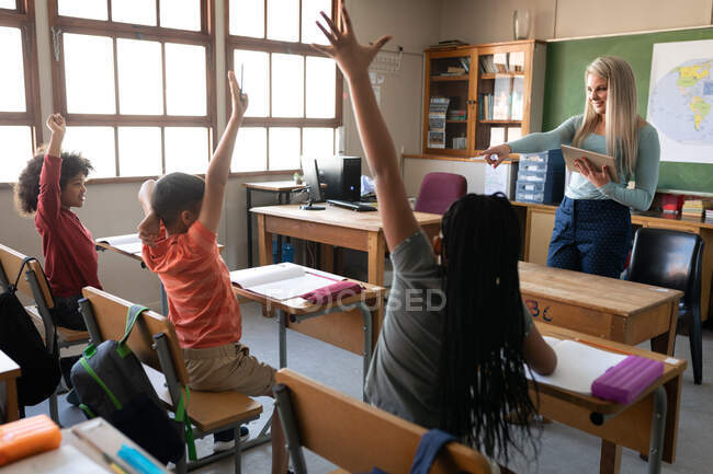 Groupe d'enfants multiethniques assis sur leur bureau pendant la leçon avec une enseignante caucasienne. Enseignement primaire distanciation sociale sécurité sanitaire pendant la pandémie de coronavirus Covid19. — Photo de stock