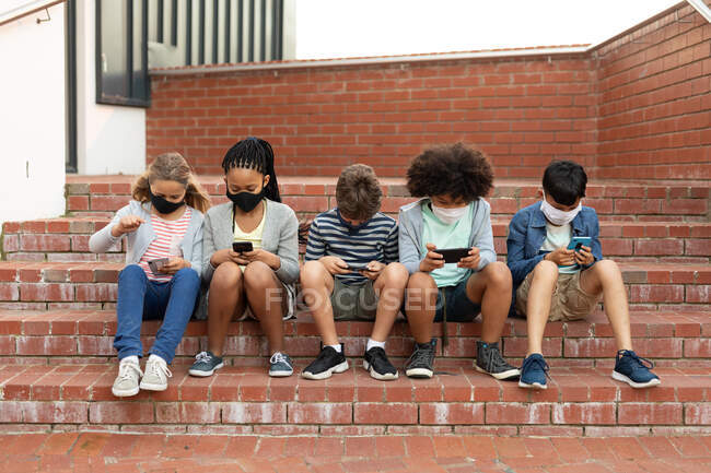 Grupo de crianças multi étnicas usando máscaras faciais usando smartphones enquanto sentado nas escadas durante uma pausa. Educação primária distanciamento social segurança sanitária durante Covid19 pandemia de coronavírus. — Fotografia de Stock