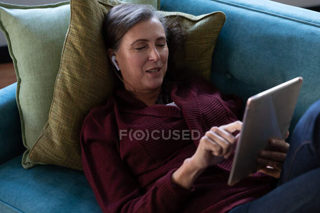 Mulher caucasiana desfrutando de tempo em casa, distanciamento social e auto-isolamento em quarentena, deitada no sofá na sala de estar, usando tablet digital e fones de ouvido sem fio. — Fotografia de Stock