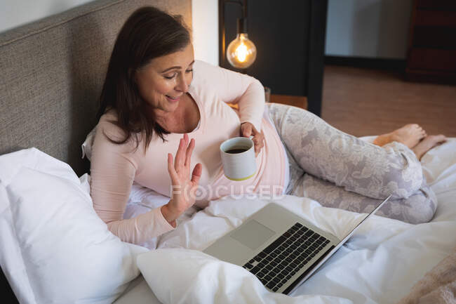 Femme caucasienne appréciant le temps à la maison, la distance sociale et l'isolement personnel en quarantaine verrouillée, couchée sur le lit dans la chambre, à l'aide d'un ordinateur portable, saluant lors d'un appel vidéo. — Photo de stock