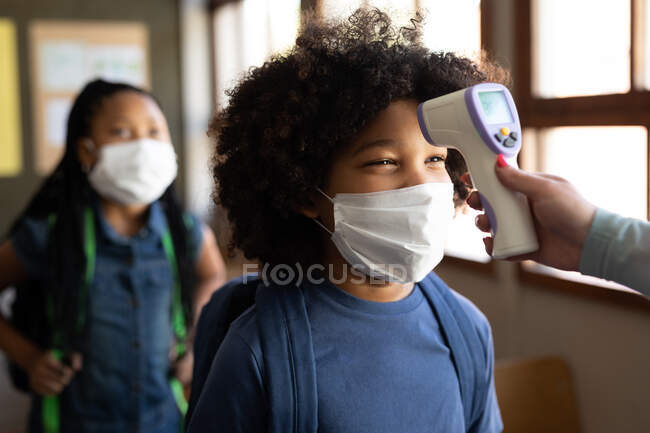 Mischlingsjunge mit Gesichtsmaske, der in einer Grundschule seine Temperatur messen lässt. Grundschulbildung soziale Distanzierung der Gesundheitssicherheit während der Covid19 Coronavirus-Pandemie. — Stockfoto