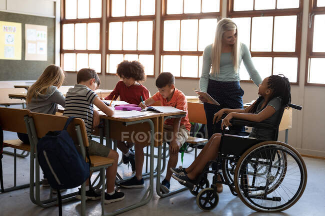 Desativar menina de raça mista sentado em sua cadeira de rodas e sua professora usando tablet na sala de aula. Educação primária distanciamento social segurança sanitária durante Covid19 pandemia de coronavírus. — Fotografia de Stock