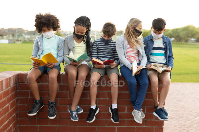Gruppe multiethnischer Kinder mit Gesichtsmasken, die Bücher lesen, während sie in einer Pause an der Wand sitzen. Grundschulbildung soziale Distanzierung der Gesundheitssicherheit während der Covid19 Coronavirus-Pandemie. — Stockfoto