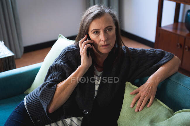 Кавказька жінка, яка проводить час удома, спілкуючись по телефону, в карантинній камері почувається ізольованою, сидить на дивані в вітальні.. — стокове фото