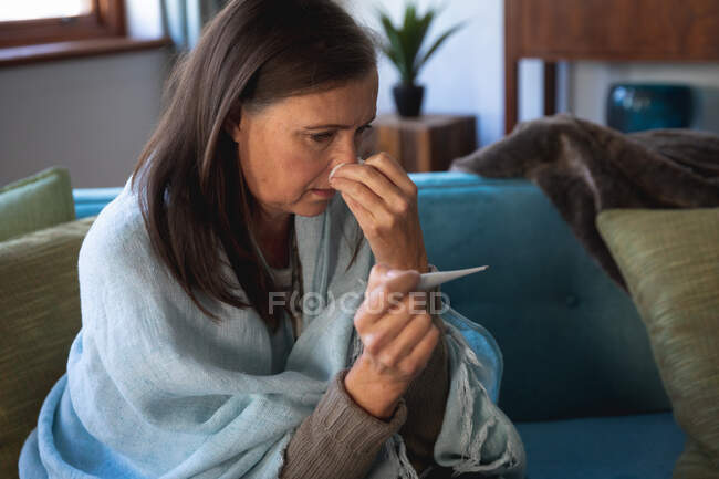 Mulher caucasiana doente passar tempo em casa, distanciamento social e auto-isolamento em quarentena bloqueio, sentado no sofá embrulhado em cobertor, segurando termômetro, medir a temperatura. — Fotografia de Stock