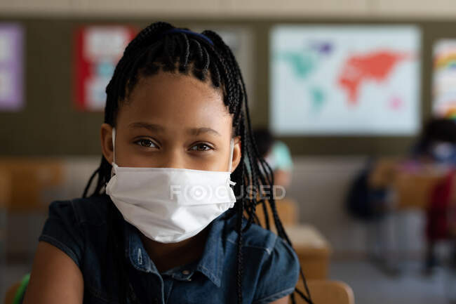 Portrait d'une fille métissée assise à un bureau portant un masque facial en classe, regardant la caméra. Enseignement primaire distanciation sociale sécurité sanitaire pendant la pandémie de coronavirus Covid19. — Photo de stock
