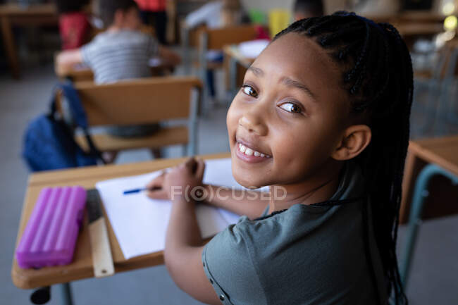 Porträt eines Mädchens mit gemischter Rasse, das auf ihrem Schreibtisch in der Schule sitzt und lächelt. Grundschulbildung soziale Distanzierung der Gesundheitssicherheit während der Covid19 Coronavirus-Pandemie. — Stockfoto