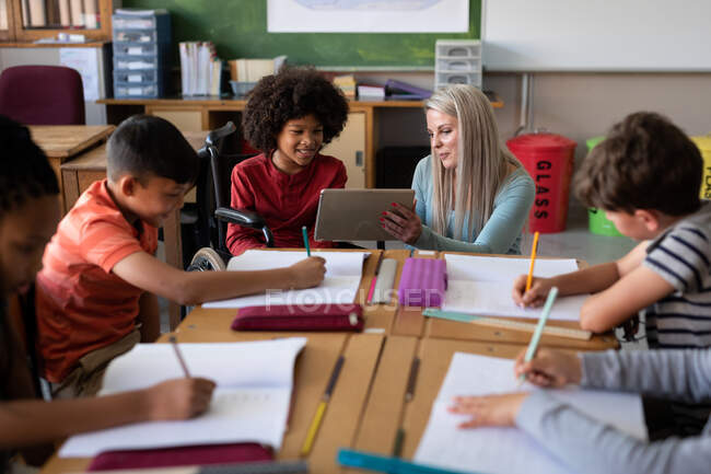 Caucasian female teacher with digital tablet teaching group of multi ethnic kids in the classroom. Primary education social distancing health safety during Covid19 Coronavirus pandemic. — Stock Photo