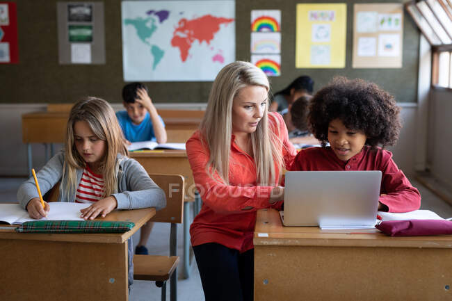 Кавказький вчитель і багатонаціональна група дітей, які користуються ноутбуком під час уроку. Первинна освіта Соціальна безпека для здоров'я під час пандемії Ковіда19 Коронавірус. — стокове фото
