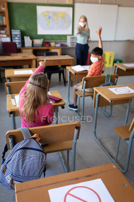 Gruppe multiethnischer Kinder, die während des Unterrichts mit einer Lehrerin mit Gesichtsmaske auf ihrem Schreibtisch sitzen. Grundschulbildung soziale Distanzierung der Gesundheitssicherheit während der Covid19 Coronavirus-Pandemie. — Stockfoto
