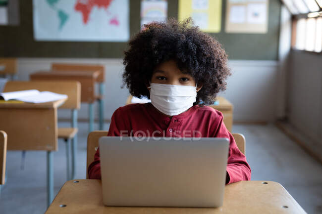 Niño de raza mixta con una máscara facial, el uso de un ordenador portátil mientras está sentado en su escritorio en la clase en la escuela. Educación primaria distanciamiento social seguridad sanitaria durante la pandemia del Coronavirus Covid19. - foto de stock
