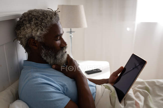 Старший афроамериканец лежит на кровати в спальне, используя цифровой планшет, потерев подбородок, социальное дистанцирование и самоизоляцию в карантинной изоляции — стоковое фото