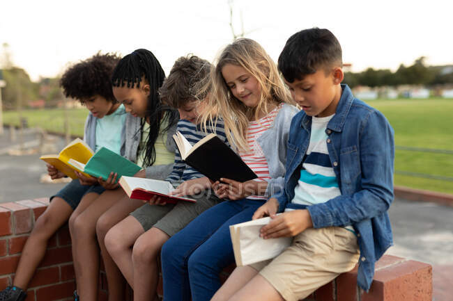 Group of multi ethnic kids reading books while sitting on the wall during a break. Primary education social distancing health safety during Covid19 Coronavirus pandemic. — Stock Photo