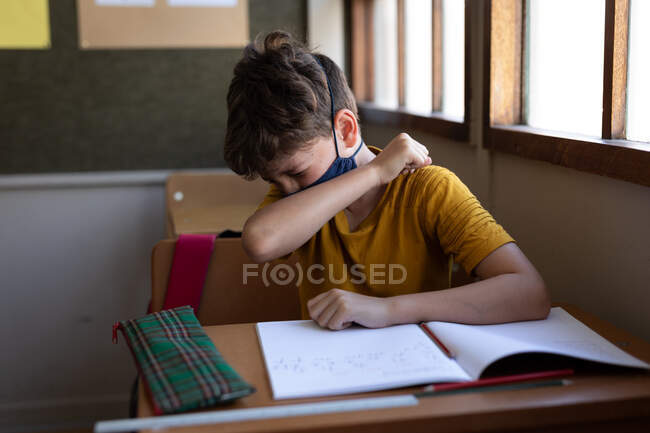 Kaukasischer Junge sitzt mit Mundschutz am Schreibtisch im Klassenzimmer und verdeckt sein Gesicht, während er niest. Grundschulbildung soziale Distanzierung der Gesundheitssicherheit während der Covid19 Coronavirus-Pandemie. — Stockfoto