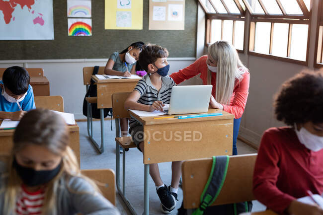 Professora caucasiana e menino caucasiano usando máscaras faciais usando laptop na escola. Educação primária distanciamento social segurança sanitária durante Covid19 pandemia de coronavírus. — Fotografia de Stock