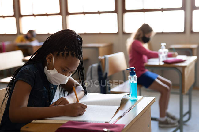 Gemischte Rassenmädchen tragen Gesichtsmaske, während sie auf ihrem Schreibtisch im Klassenzimmer mit einem Desinfektionsmittel sitzen. Grundschulbildung soziale Distanzierung der Gesundheitssicherheit während der Covid19 Coronavirus-Pandemie. — Stockfoto