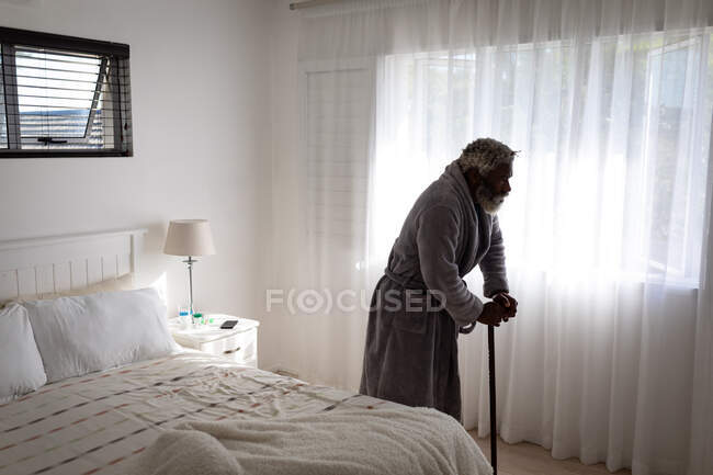 Hombre mayor afroamericano caminando con un bastón en un dormitorio, distanciamiento social y aislamiento en cuarentena - foto de stock