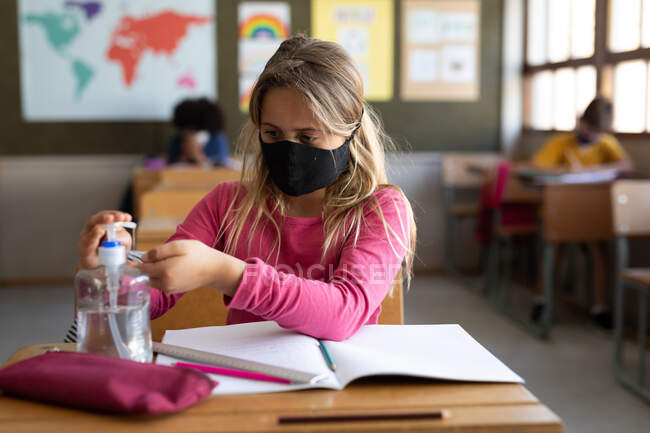 Белая девушка в маске, сидя за столом и дезинфицируя руки. Начальное образование Социальное дистанцирование безопасности здоровья во время пандемии Coronavirus Covid19. — стоковое фото