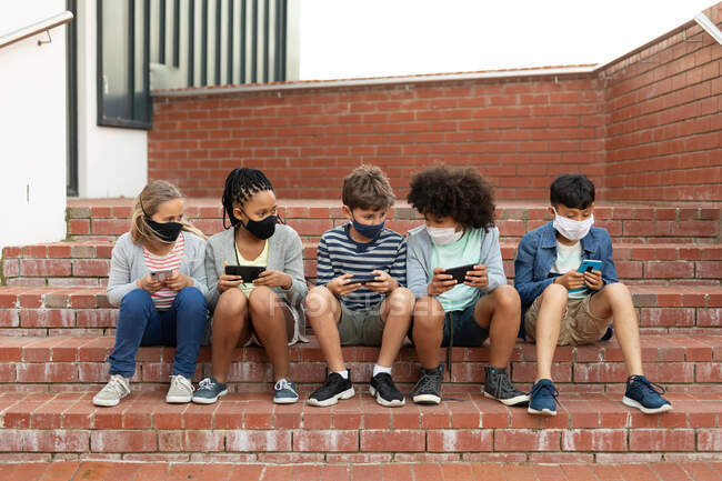 Grupo de niños multiétnicos que usan máscaras faciales usando teléfonos inteligentes mientras están sentados en las escaleras durante un descanso. Educación primaria distanciamiento social seguridad sanitaria durante la pandemia del Coronavirus Covid19. - foto de stock