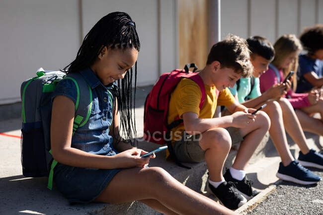 Grupo multiétnico de niños de la escuela primaria que usan teléfonos inteligentes mientras están sentados juntos. Educación primaria distanciamiento social seguridad sanitaria durante la pandemia del Coronavirus Covid19 - foto de stock