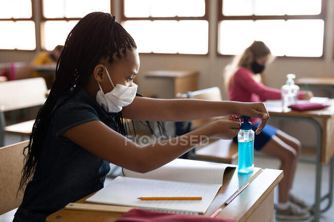 Chica de raza mixta que usa mascarilla facial, desinfectando sus manos mientras está sentada en su escritorio en el aula. Educación primaria distanciamiento social seguridad sanitaria durante la pandemia del Coronavirus Covid19. - foto de stock