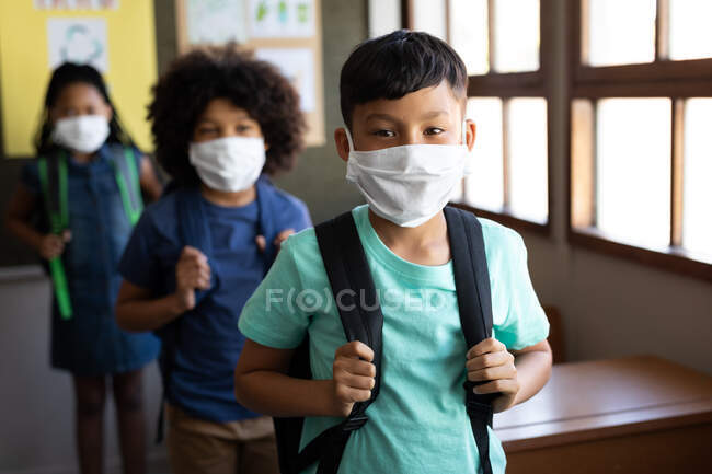 Grupo multiétnico de niños de la escuela primaria mirando a la cámara, con máscaras faciales en el salón de la escuela. Educación primaria distanciamiento social seguridad sanitaria durante la pandemia del Coronavirus Covid19. - foto de stock