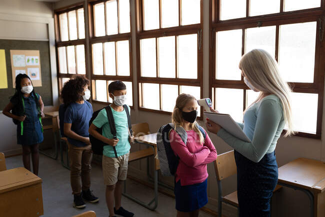 Caucasian female teacher wearing face mask measuring temperature of children in an elementary school. Primary education social distancing health safety during Covid19 Coronavirus pandemic. — Stock Photo