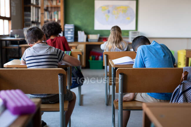Vue arrière d'un groupe d'enfants multiethniques étudiant assis sur leur bureau à l'école. Enseignement primaire distanciation sociale sécurité sanitaire pendant la pandémie de coronavirus Covid19. — Photo de stock