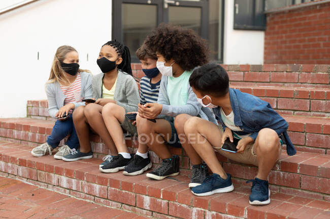 Grupo de niños multiétnicos que usan máscaras faciales usando teléfonos inteligentes mientras están sentados en las escaleras durante un descanso. Educación primaria distanciamiento social seguridad sanitaria durante la pandemia del Coronavirus Covid19. - foto de stock