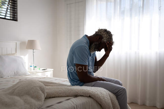 African American senior man sitting on a bed in a bedroom, holding his forehead in his hand, social distancing and self isolation in quarantine lockdown — Stock Photo
