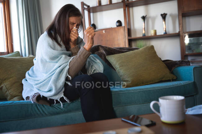 Хвора кавказька жінка проводить вдома час, дистанціюючись і самоізолюючись в карантині, сидячи на дивані загорненій в ковдру, чхаючи.. — стокове фото